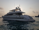 A large white yacht is floating on calm waters during what appears to be either sunrise or sunset. The yacht has a sleek design with antennas and satellite dishes visible on the top. The sky is slightly overcast with clouds, and the water appears reflective with a gentle movement.