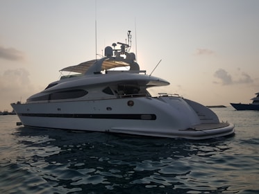 A large white yacht is floating on calm waters during what appears to be either sunrise or sunset. The yacht has a sleek design with antennas and satellite dishes visible on the top. The sky is slightly overcast with clouds, and the water appears reflective with a gentle movement.