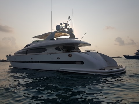 A large white yacht is floating on calm waters during what appears to be either sunrise or sunset. The yacht has a sleek design with antennas and satellite dishes visible on the top. The sky is slightly overcast with clouds, and the water appears reflective with a gentle movement.