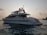 A large white yacht is floating on calm waters during what appears to be either sunrise or sunset. The yacht has a sleek design with antennas and satellite dishes visible on the top. The sky is slightly overcast with clouds, and the water appears reflective with a gentle movement.