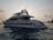 A large white yacht is floating on calm waters during what appears to be either sunrise or sunset. The yacht has a sleek design with antennas and satellite dishes visible on the top. The sky is slightly overcast with clouds, and the water appears reflective with a gentle movement.