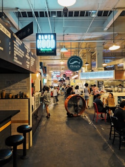 A bustling indoor market scene with several food stalls. Signs for 'Ramen Hood' and 'Prawn Coastal' hang prominently above. People are walking through the corridor, some are standing in line, and others are sitting at tables enjoying meals. The lighting is warm and industrial, with exposed light fixtures and a high ceiling.