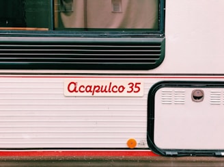 The image features the side of what appears to be a vehicle or trailer with the name 'Acapulco 35' written in red on a white background. There are horizontal ridges, black detailing, and a small, round, orange reflector mounted on the lower section.