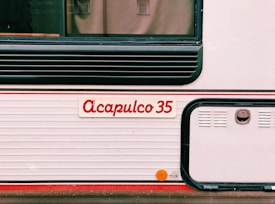 The image features the side of what appears to be a vehicle or trailer with the name 'Acapulco 35' written in red on a white background. There are horizontal ridges, black detailing, and a small, round, orange reflector mounted on the lower section.
