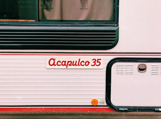 The image features the side of what appears to be a vehicle or trailer with the name 'Acapulco 35' written in red on a white background. There are horizontal ridges, black detailing, and a small, round, orange reflector mounted on the lower section.