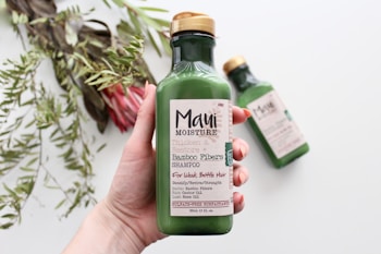 A hand holding a green bottle of shampoo labeled 'Maui Moisture' with bamboo fibers, intended for thickening and restoring hair. In the background, another identical bottle is slightly out of focus on a white surface. To the left, there are green leafy plants and a pink flower slightly blurred.