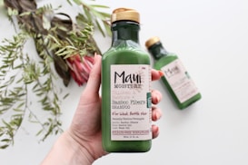 A hand holding a green bottle of shampoo labeled 'Maui Moisture' with bamboo fibers, intended for thickening and restoring hair. In the background, another identical bottle is slightly out of focus on a white surface. To the left, there are green leafy plants and a pink flower slightly blurred.