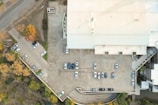 Aerial view of a parking lot adjacent to a large industrial or commercial building. Several vehicles are parked in designated spaces, with some areas showing more concentration of cars than others. The building has a light-colored roof and is surrounded by trees with autumn foliage, creating a contrast between the urban structure and natural elements.