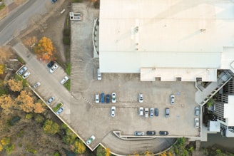 Aerial view of a parking lot adjacent to a large industrial or commercial building. Several vehicles are parked in designated spaces, with some areas showing more concentration of cars than others. The building has a light-colored roof and is surrounded by trees with autumn foliage, creating a contrast between the urban structure and natural elements.