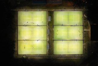 Aerial view of six illuminated tennis courts arranged in two rows of three. The courts are surrounded by darkness and a few scattered objects are visible on and around the courts. Some courts have players present while others are empty.