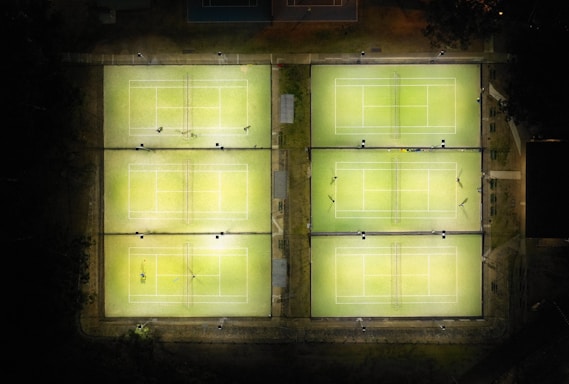 Aerial view of six illuminated tennis courts arranged in two rows of three. The courts are surrounded by darkness and a few scattered objects are visible on and around the courts. Some courts have players present while others are empty.