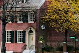 A charming brick house with dark green window shutters and a cream-colored door adorned with a wreath. The scene is set in autumn, with a tree on the right displaying yellow leaves. A classic black lamppost stands in front of the house, and green street signs indicate the intersection of High St. and another road.