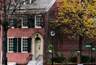 A charming brick house with dark green window shutters and a cream-colored door adorned with a wreath. The scene is set in autumn, with a tree on the right displaying yellow leaves. A classic black lamppost stands in front of the house, and green street signs indicate the intersection of High St. and another road.