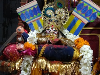 An ornate, traditional mask with elaborate designs and bright colors is surrounded by vibrant garlands. The mask features striking eyes and intricate embellishments, including a peacock feather on top. The backdrop includes bold patterns in blue, yellow, and green.