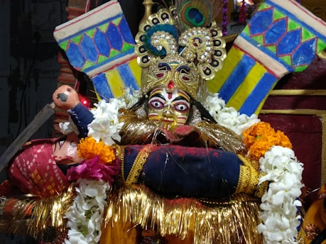 An ornate, traditional mask with elaborate designs and bright colors is surrounded by vibrant garlands. The mask features striking eyes and intricate embellishments, including a peacock feather on top. The backdrop includes bold patterns in blue, yellow, and green.