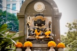 A detailed sculpture of a multi-armed deity is adorned with bright yellow marigold garlands. The statue is placed within a stone arch structure, featuring intricate carvings and a prominent Om symbol. Lush green foliage surrounds the area, complemented by a modern building visible in the background.