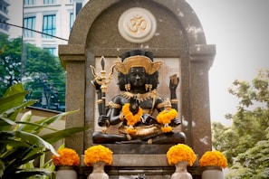 A detailed sculpture of a multi-armed deity is adorned with bright yellow marigold garlands. The statue is placed within a stone arch structure, featuring intricate carvings and a prominent Om symbol. Lush green foliage surrounds the area, complemented by a modern building visible in the background.