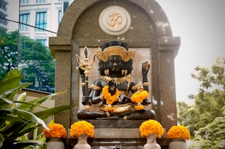 A detailed sculpture of a multi-armed deity is adorned with bright yellow marigold garlands. The statue is placed within a stone arch structure, featuring intricate carvings and a prominent Om symbol. Lush green foliage surrounds the area, complemented by a modern building visible in the background.
