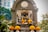 A detailed sculpture of a multi-armed deity is adorned with bright yellow marigold garlands. The statue is placed within a stone arch structure, featuring intricate carvings and a prominent Om symbol. Lush green foliage surrounds the area, complemented by a modern building visible in the background.