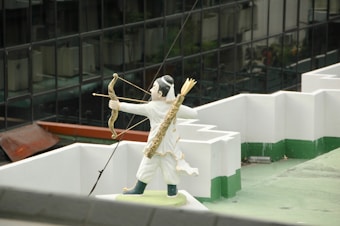 A statue of an archer in white and gold attire is positioned on a rooftop, drawing a bow with an arrow ready to shoot. The figure carries a quiver filled with arrows on its back. In the background, there are reflective glass windows of a tall building.
