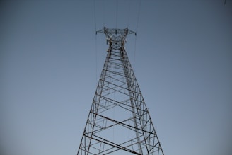 A tall metal transmission tower rises against a clear sky, with power lines extending outward from the structure.