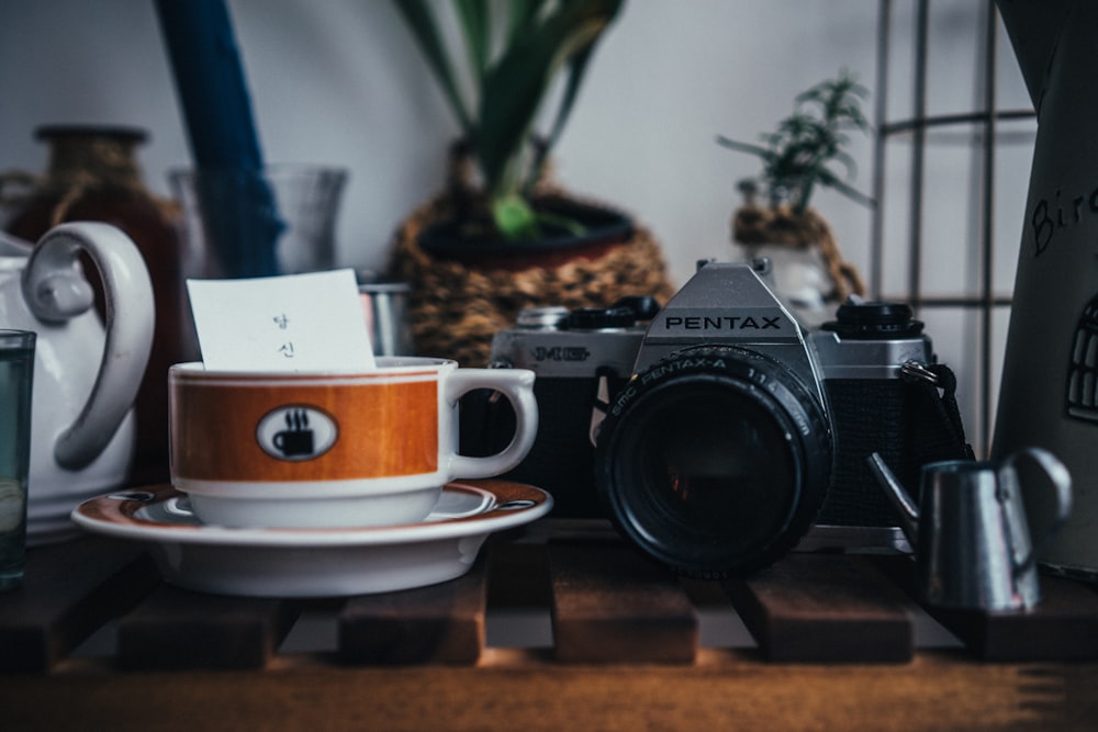 gray and black Pentax MILC camera beside round white and brown ceramic teacup on top of round white and brown saucer on top of table