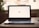 A laptop displaying the word 'Hi' on its screen is placed on a wooden table with a soft background blur. To the right, a pair of earphones is partially visible.