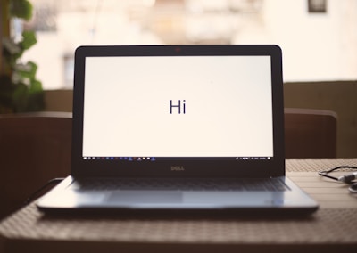 A laptop displaying the word 'Hi' on its screen is placed on a wooden table with a soft background blur. To the right, a pair of earphones is partially visible.