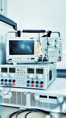 A stack of electronic measurement and testing equipment including an oscilloscope and a programmable power supply. The oscilloscope has a screen and various dials and buttons, while the power supply displays digital readings and has multiple colored cables connected. The setup suggests a technical or laboratory environment.