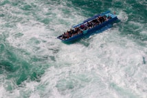 A small tour boat filled with people navigates through turbulent, foamy waters. The boat is blue and has a sign indicating a short tour. The water around is a mix of green and white due to the churning of waves.