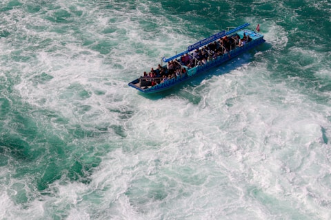 A small tour boat filled with people navigates through turbulent, foamy waters. The boat is blue and has a sign indicating a short tour. The water around is a mix of green and white due to the churning of waves.