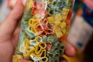 A close-up view of a hand holding a clear plastic package containing colorful pasta in various shapes. The pasta is in shades of yellow, red, and green.