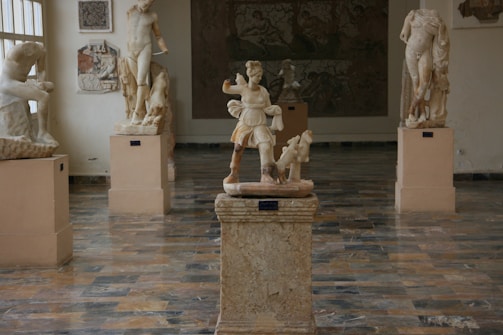 A gallery with various classical sculptures displayed on pedestals. The floor is made of polished stone tiles, reflecting the soft natural light coming in from the window. The sculptures depict figures in different poses, with one central statue prominently set on a pedestal in the middle of the room.