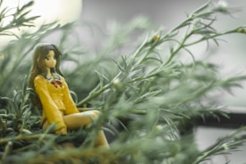 A figurine of an anime character dressed in a yellow outfit with long brown hair sits among dense, green, leafy plants. The background is softly blurred, creating a peaceful and serene atmosphere.