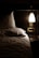 A cozy, dimly lit bedroom features a neatly made bed with white pillows and sheets. A small bedside table holds a few items, illuminated by a modern wall-mounted lamp casting a warm glow. The setting suggests a quiet, intimate atmosphere suited for relaxation and rest.