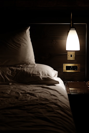 A cozy, dimly lit bedroom features a neatly made bed with white pillows and sheets. A small bedside table holds a few items, illuminated by a modern wall-mounted lamp casting a warm glow. The setting suggests a quiet, intimate atmosphere suited for relaxation and rest.