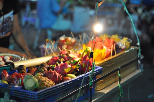 A vibrant street market stall displays an array of colorful tropical fruits, including dragon fruit, mango, and rambutan. These fruits are arranged in a blue crate and are illuminated by a bright overhead bulb, casting lively shadows and highlights. In the background, a vendor tends to the stall while blurred people are seated, creating a bustling yet cozy nighttime ambiance.
