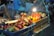 A vibrant street market stall displays an array of colorful tropical fruits, including dragon fruit, mango, and rambutan. These fruits are arranged in a blue crate and are illuminated by a bright overhead bulb, casting lively shadows and highlights. In the background, a vendor tends to the stall while blurred people are seated, creating a bustling yet cozy nighttime ambiance.