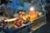 A vibrant street market stall displays an array of colorful tropical fruits, including dragon fruit, mango, and rambutan. These fruits are arranged in a blue crate and are illuminated by a bright overhead bulb, casting lively shadows and highlights. In the background, a vendor tends to the stall while blurred people are seated, creating a bustling yet cozy nighttime ambiance.