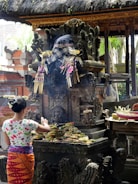 A person is making offerings at a traditional Balinese shrine, which features intricate stone carvings and is adorned with checkered cloth and floral decorations. The scene is set in an outdoor temple environment with thatched roofs and surrounding greenery.