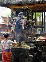 A person is making offerings at a traditional Balinese shrine, which features intricate stone carvings and is adorned with checkered cloth and floral decorations. The scene is set in an outdoor temple environment with thatched roofs and surrounding greenery.