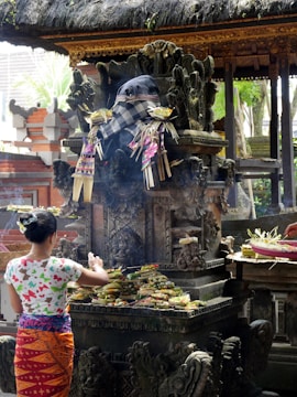 A person is making offerings at a traditional Balinese shrine, which features intricate stone carvings and is adorned with checkered cloth and floral decorations. The scene is set in an outdoor temple environment with thatched roofs and surrounding greenery.