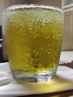 A glass filled with a yellow, fizzy liquid is placed on a folded white napkin. The liquid is carbonated, with bubbles visibly clinging to the sides of the glass. The background appears to be indoors, possibly a living room or office, with neutral tones and an air conditioner vent visible.