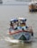 A small, colorful fishing boat navigates through the water with two people aboard. Both are standing at the front, one holding onto the railing. The boat shows signs of frequent use, evident from its weathered appearance. In the background, other larger boats are visible, suggesting a busy harbor area.