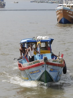 A small, colorful fishing boat navigates through the water with two people aboard. Both are standing at the front, one holding onto the railing. The boat shows signs of frequent use, evident from its weathered appearance. In the background, other larger boats are visible, suggesting a busy harbor area.