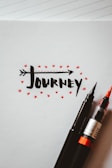 The artwork features the word 'journey' written in a bold, black, stylized font with an arrow integrated into the text. Surrounding the text are small red stars, adding a decorative touch. Two pens, one with a black tip and the other with a red brush tip, rest on the bottom right corner of the paper. The background is a blank notebook page with faint lines visible.