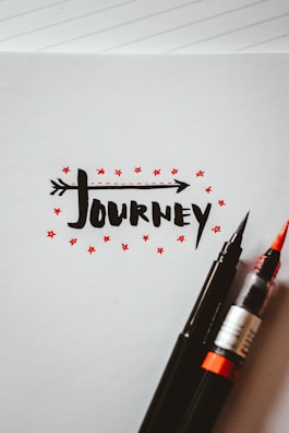 The artwork features the word 'journey' written in a bold, black, stylized font with an arrow integrated into the text. Surrounding the text are small red stars, adding a decorative touch. Two pens, one with a black tip and the other with a red brush tip, rest on the bottom right corner of the paper. The background is a blank notebook page with faint lines visible.