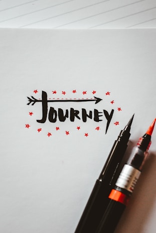 The artwork features the word 'journey' written in a bold, black, stylized font with an arrow integrated into the text. Surrounding the text are small red stars, adding a decorative touch. Two pens, one with a black tip and the other with a red brush tip, rest on the bottom right corner of the paper. The background is a blank notebook page with faint lines visible.