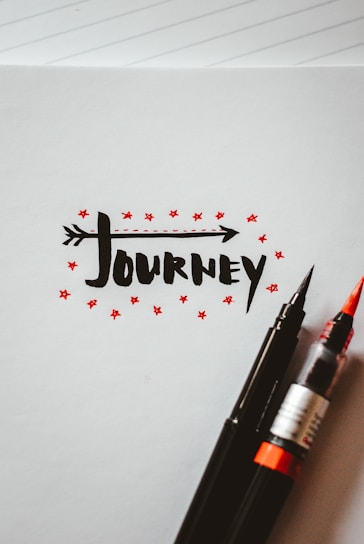 The artwork features the word 'journey' written in a bold, black, stylized font with an arrow integrated into the text. Surrounding the text are small red stars, adding a decorative touch. Two pens, one with a black tip and the other with a red brush tip, rest on the bottom right corner of the paper. The background is a blank notebook page with faint lines visible.