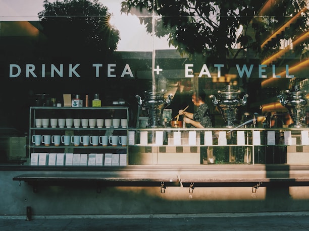 A café storefront with large glass windows displaying the phrase 'DRINK TEA + EAT WELL'. Inside, shelves hold numerous neatly arranged white cups and mugs, while a person is working with tea or coffee-making equipment. The sun casts long shadows and creates a warm glow inside the shop, adding to a cozy atmosphere.